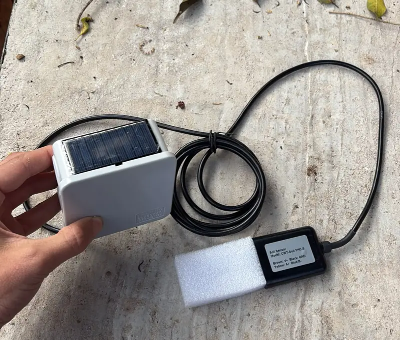 A NuaSense node box with its small solar panel on top, held in one hand, and the soil sensor on its cable in front of it